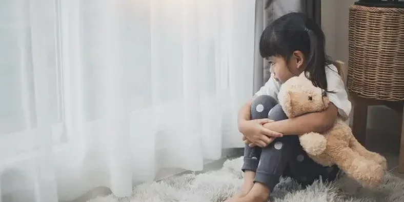 anak kecil sedih sambil memegang boneka dan melihat ke arah jendela