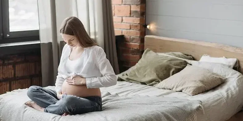 wanita hamil duduk di atas kasur sambil memegang perutnya